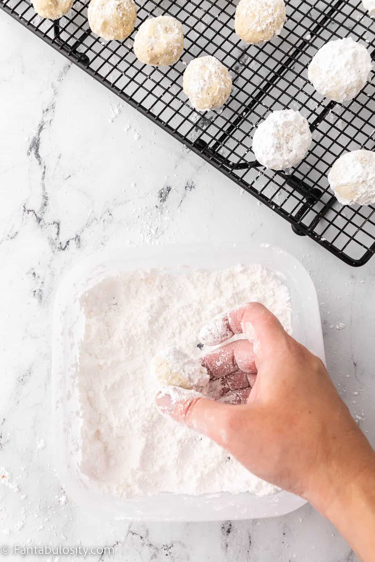 A hand holding a pecan snowball cookie is rolling it in powdered sugar while a wire rack in the background holds additional cookies that are in various stages of coating