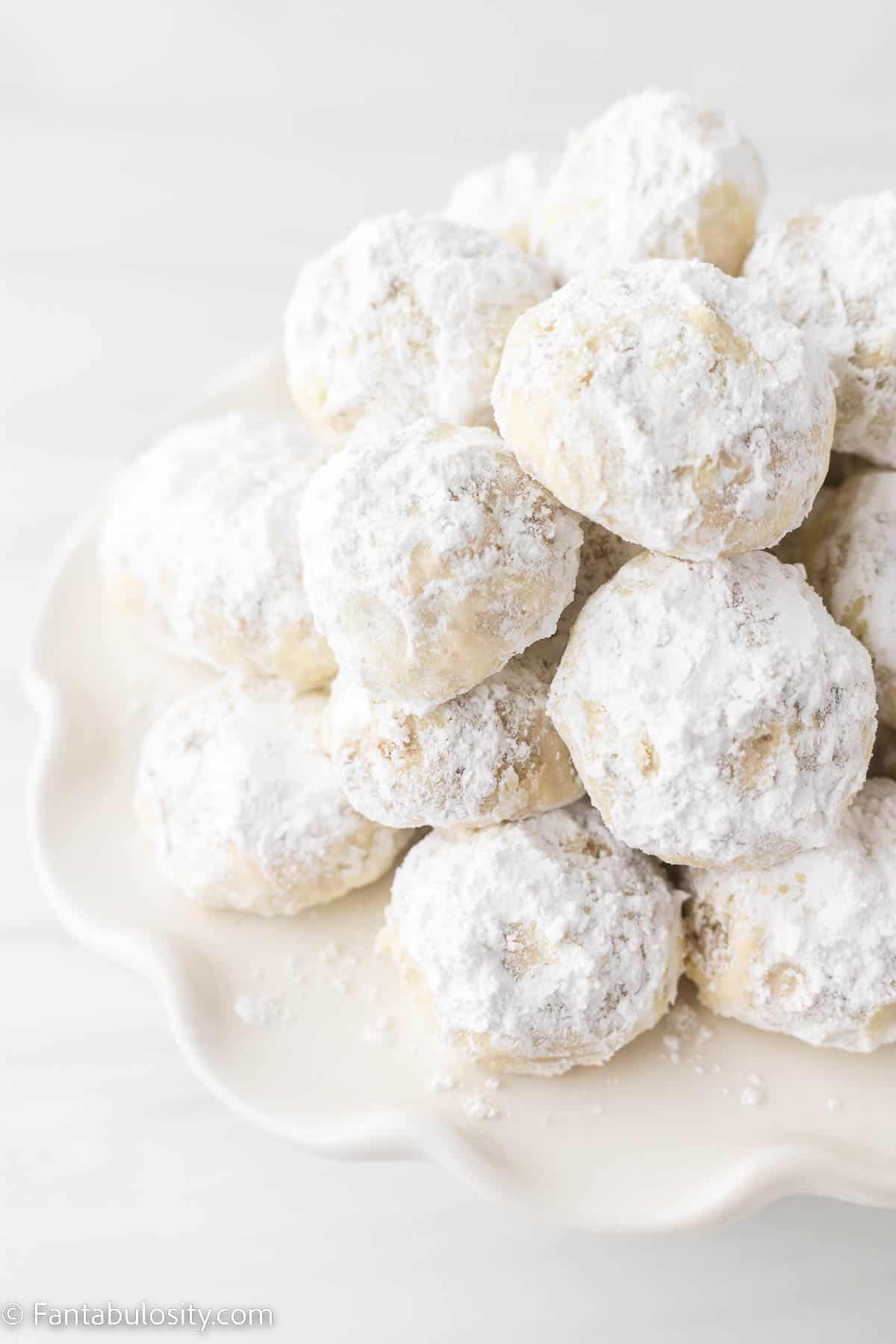 A close of photo of powdered sugar coated snowball cookies piled on a decorative white cake stand