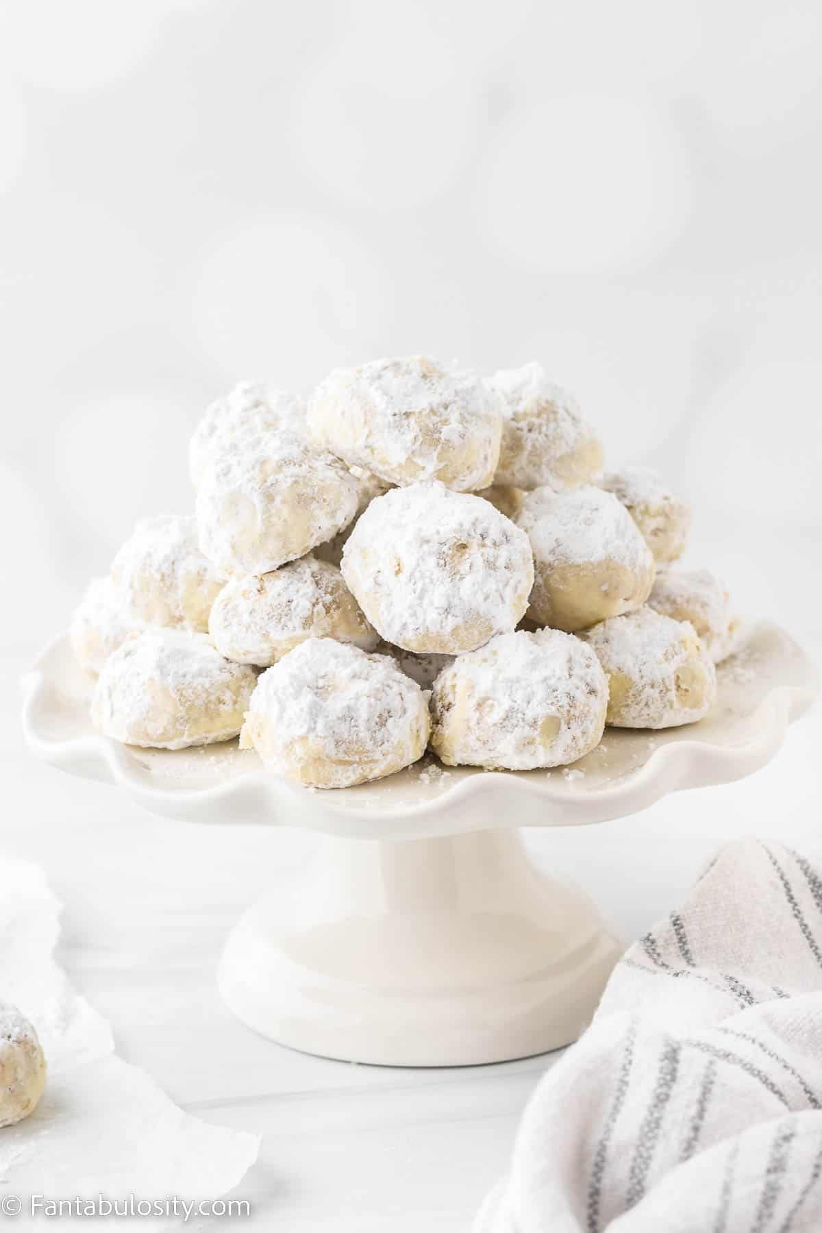 Powdered sugar coated pecan snowball cookies are piled on a white cake stand