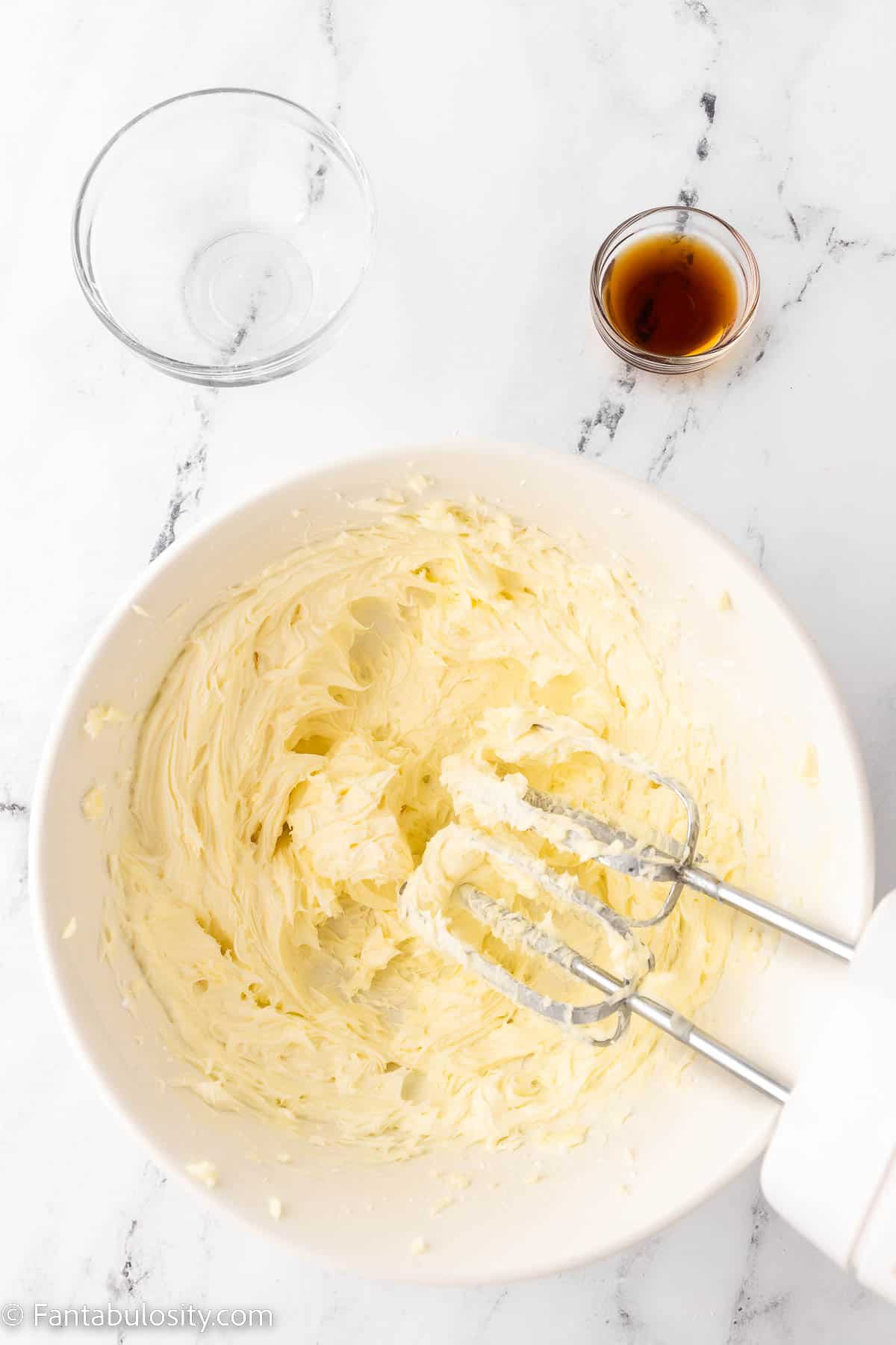 Two sticks of butter, vanilla extract and powdered sugar have been mixed with a hand mixer in a large mixing bowl until they are fluffy and light yellow
