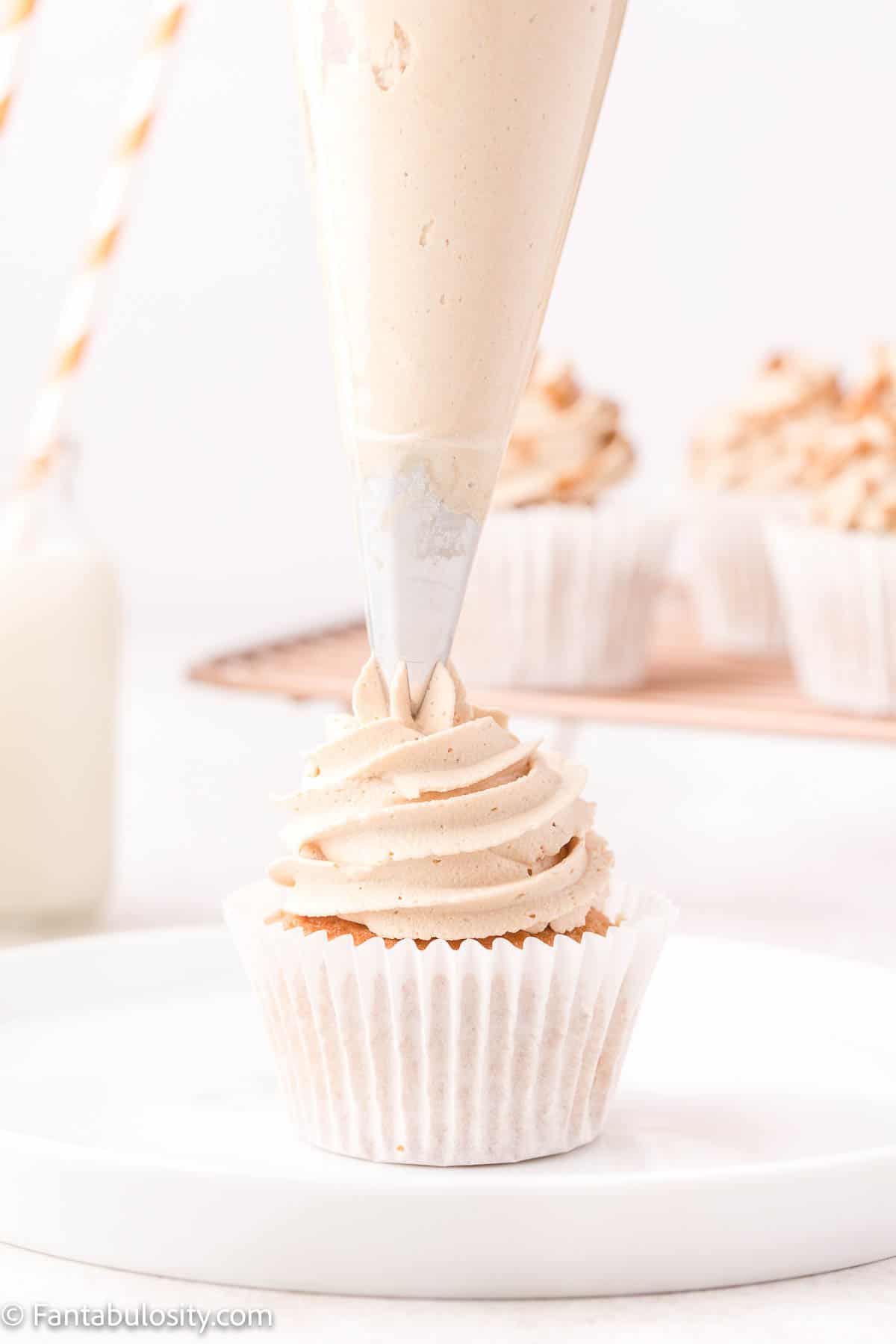A single cupcake is shown on a white plate and a piping bag of frosting is visible piping frosting onto the cupcake