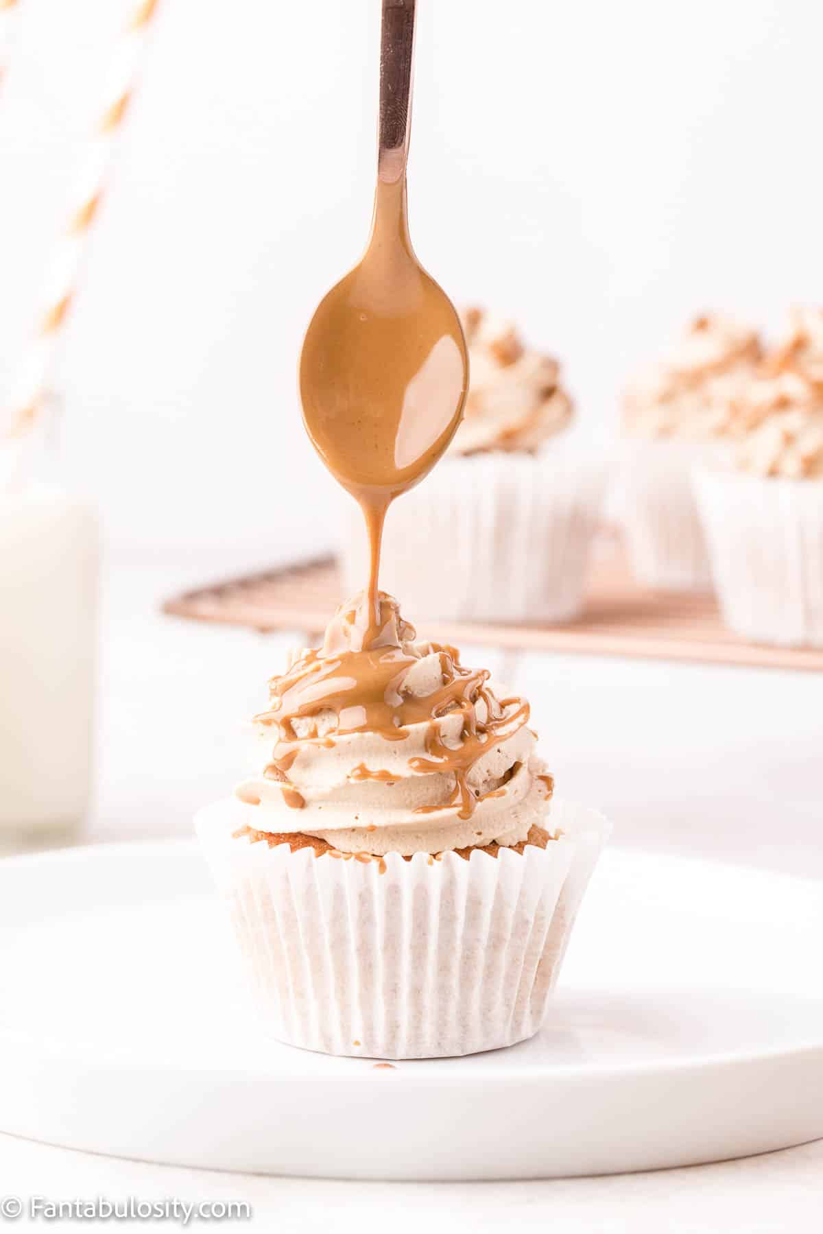 A spoon with melted cookie butter is drizzling the Biscoff spread over a frosted cupcake