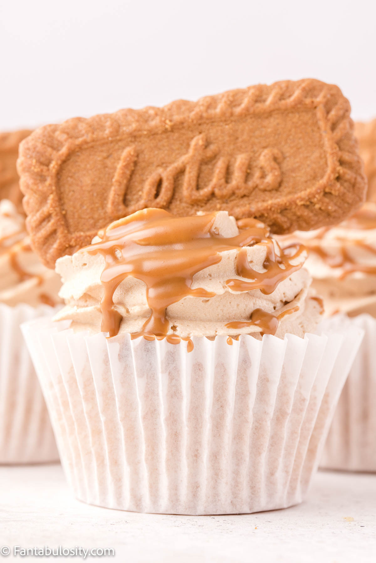 A frosted cupcake topped with buttercream, a drizzle of Biscoff spread and a Biscoff cookie is centered with additional cupcakes visible in the background