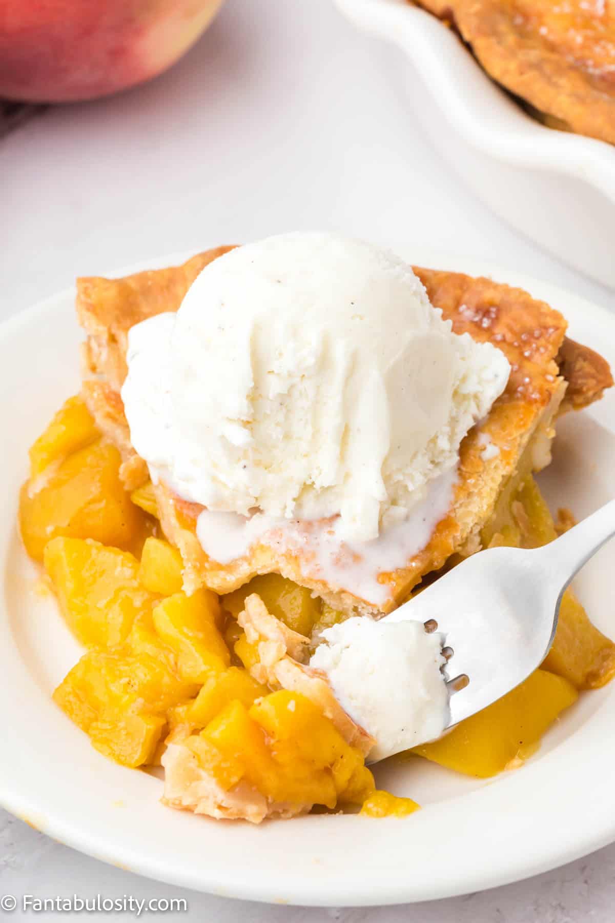 A slice of peach mango pie with vanilla ice cream on top. A fork is taking a piece from the pie piece. 
