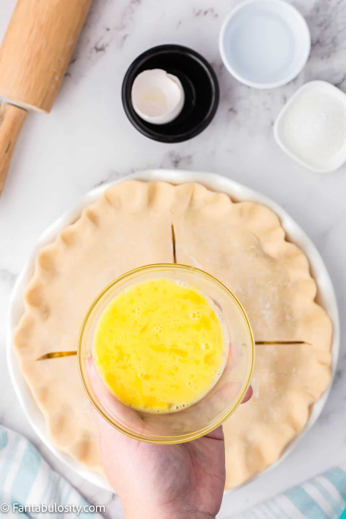 A bowl of egg wash being held above an unbaked pie.