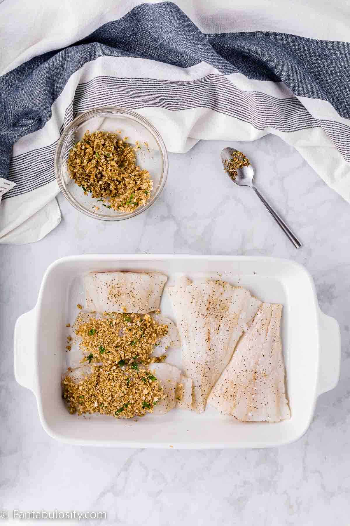 Raw cod filets in baking dish with panko and seasonings being added on top.