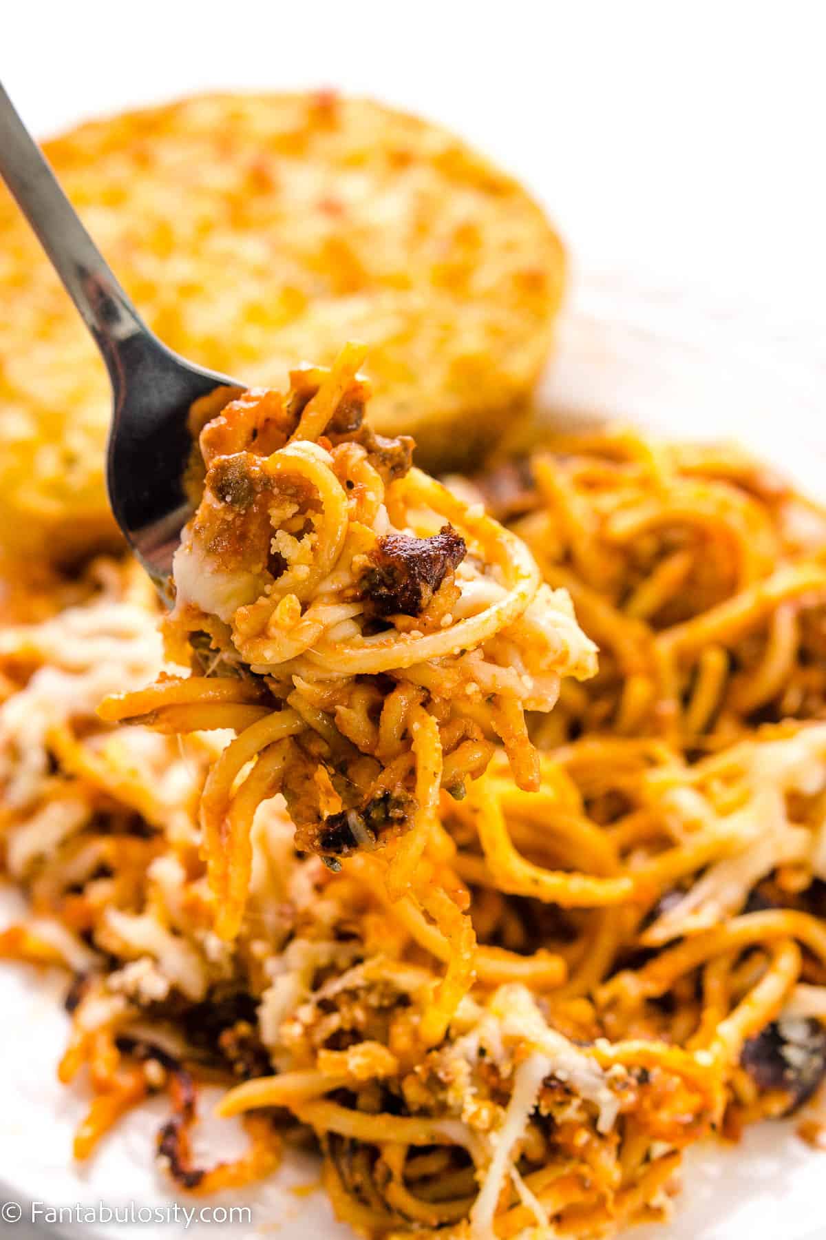 A fork holding a bite of fried spaghetti, over a plate of it, next to a piece of garlic toast.