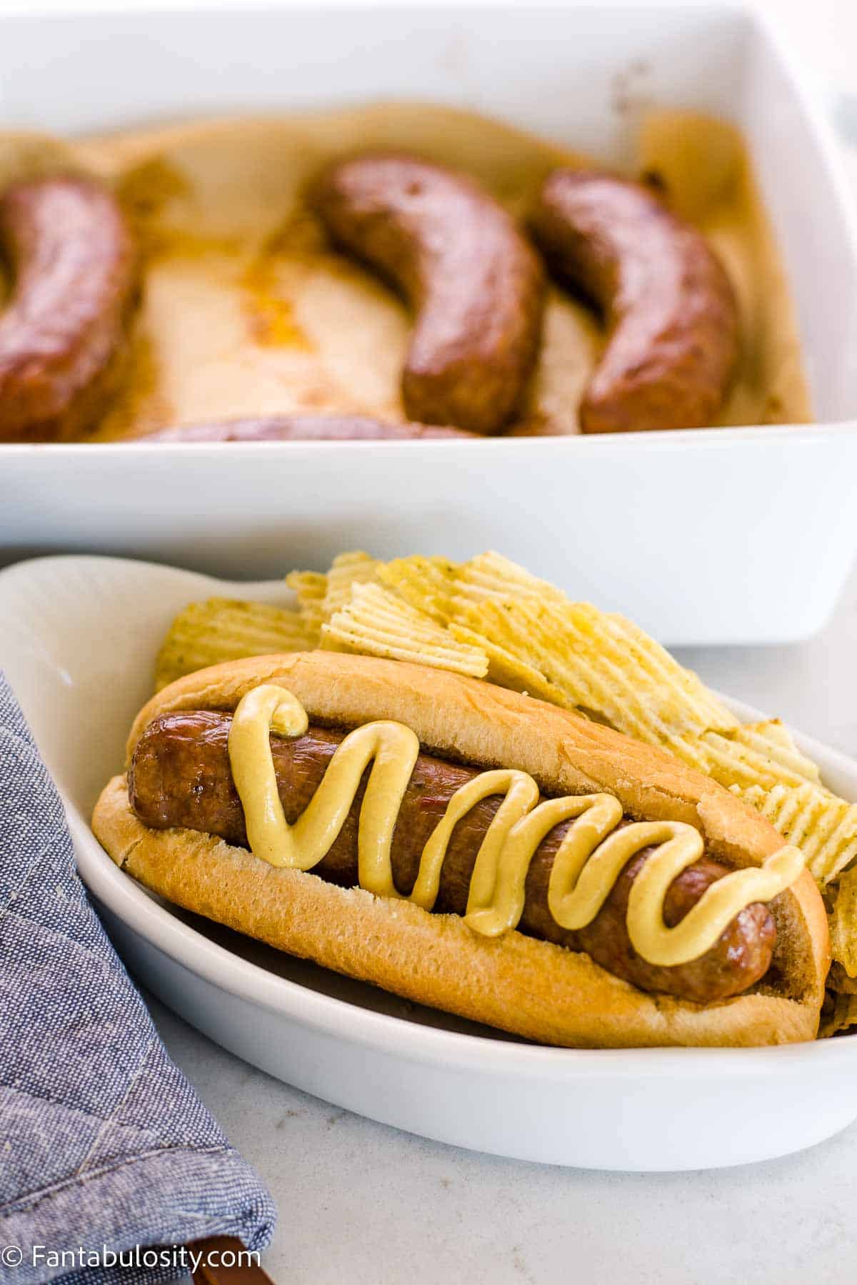 Cooked Italian sausage in bun, in a white dish next to potato chips.