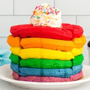 Rainbow waffles stacked on a white plate, with whipped cream on top.