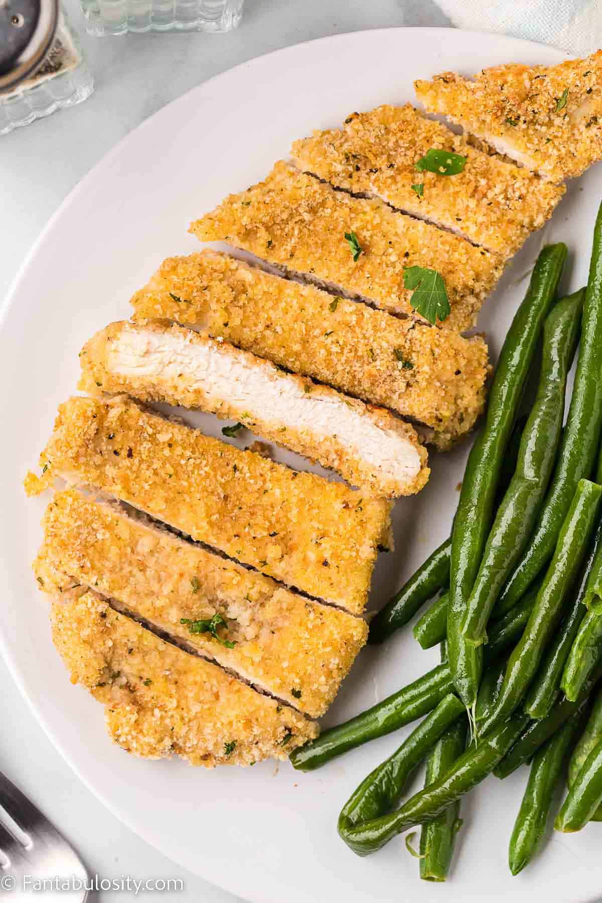 Baked chicken cutlet on white plate sitting next to green beans.