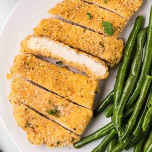 Sliced baked chicken cutlets on white plate, next to green beans.