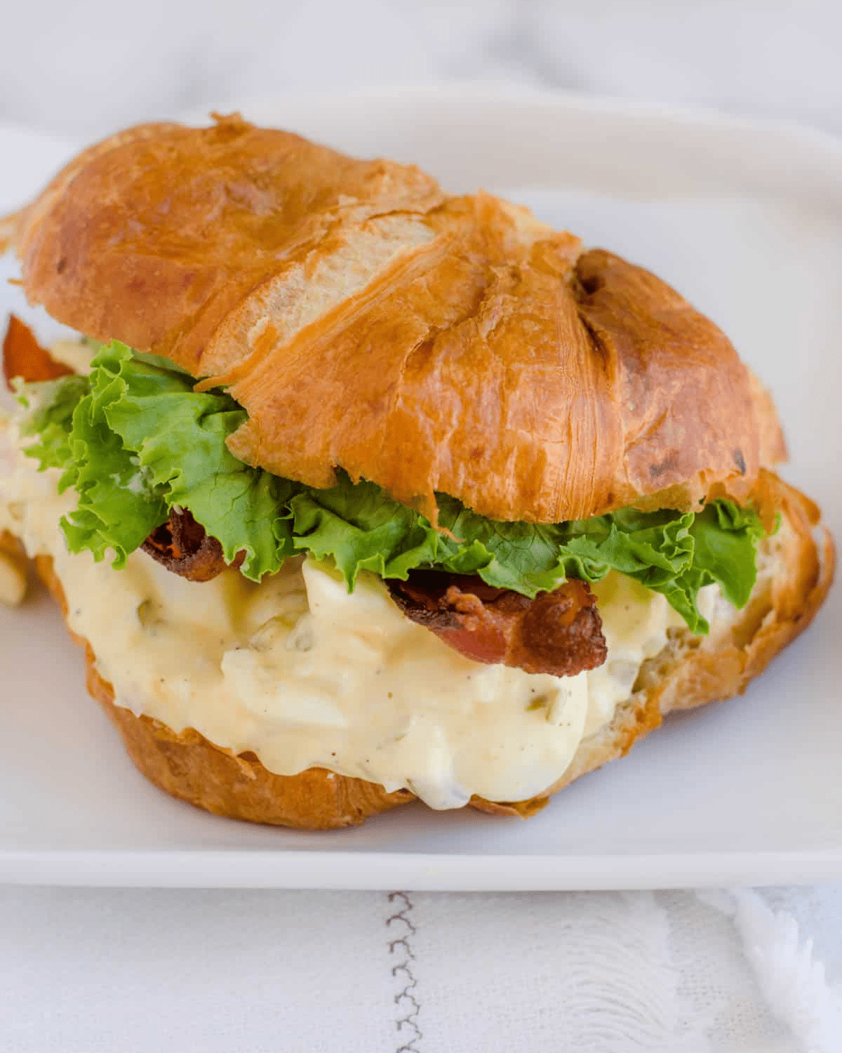 Egg salad on a croissant with lettuce and bacon.