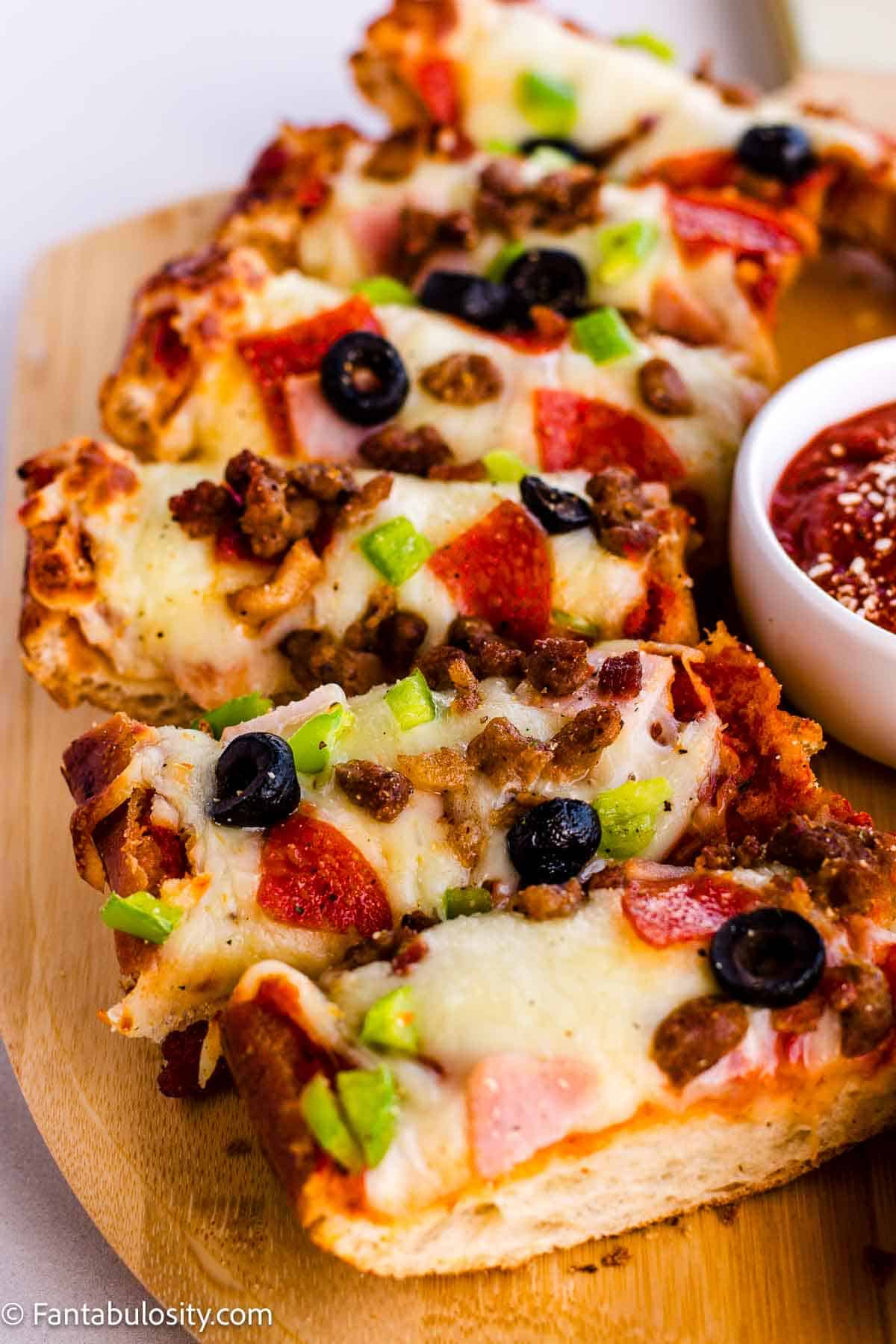Sliced French bread pizza slices on wooden cutting board with meat and vegetable toppings.