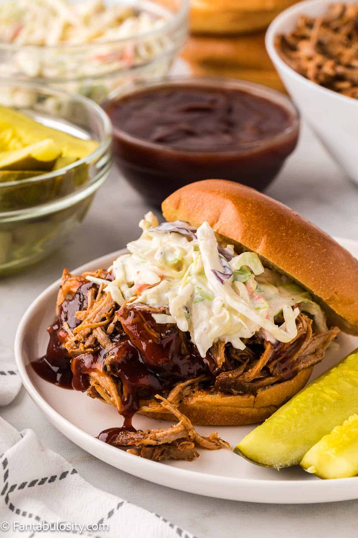 Shredded pork butt on bun, with coleslaw on top, and sitting on a white plate.