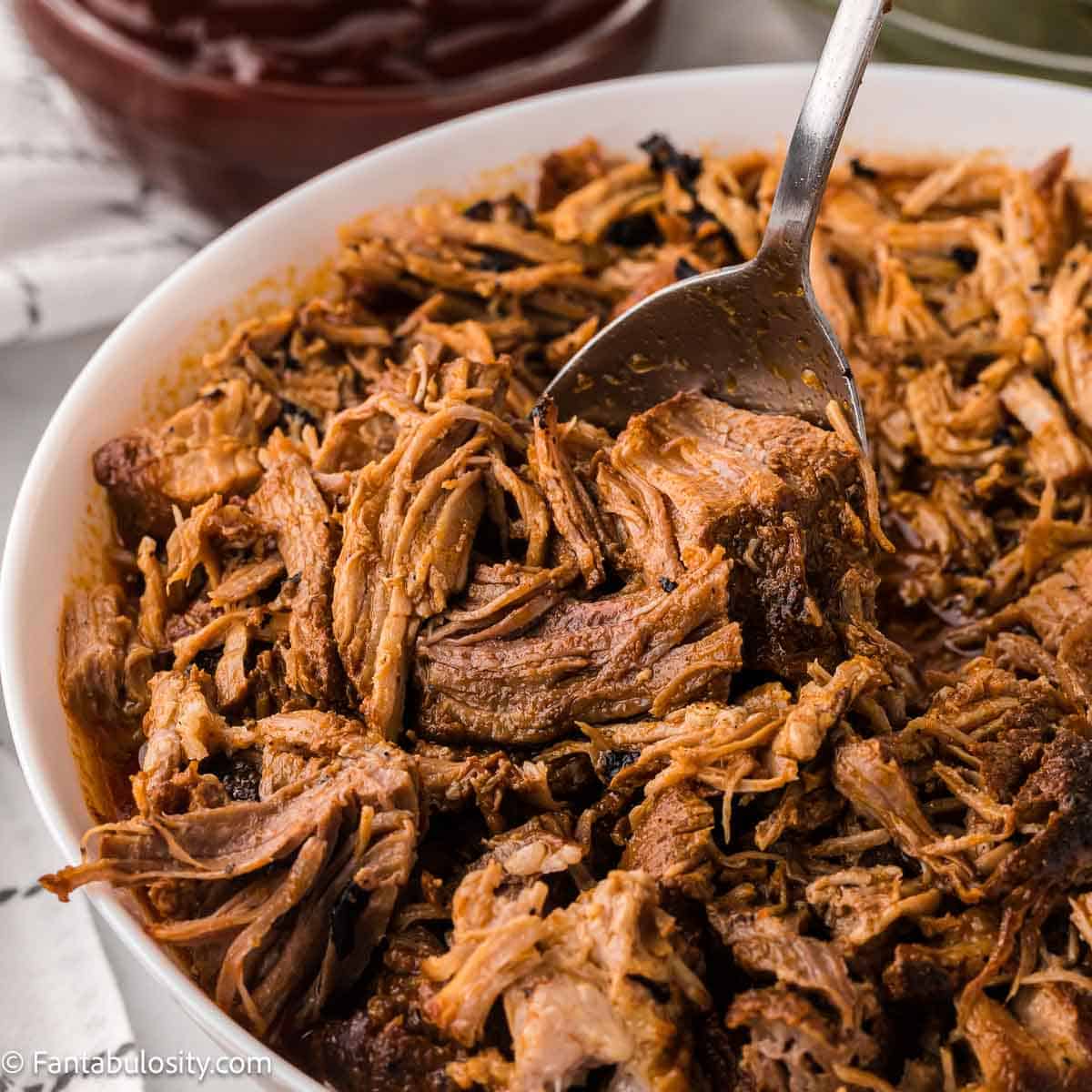 Cooked Instant Pot pork butt in white bowl, sitting next to barbecue sauce.