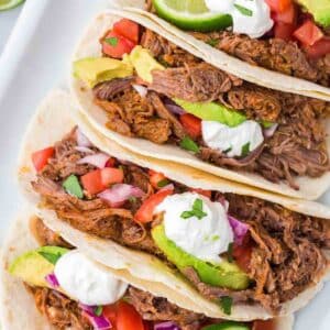 Shredded beef tacos that were cooked in slow cooker, sitting on white plate.