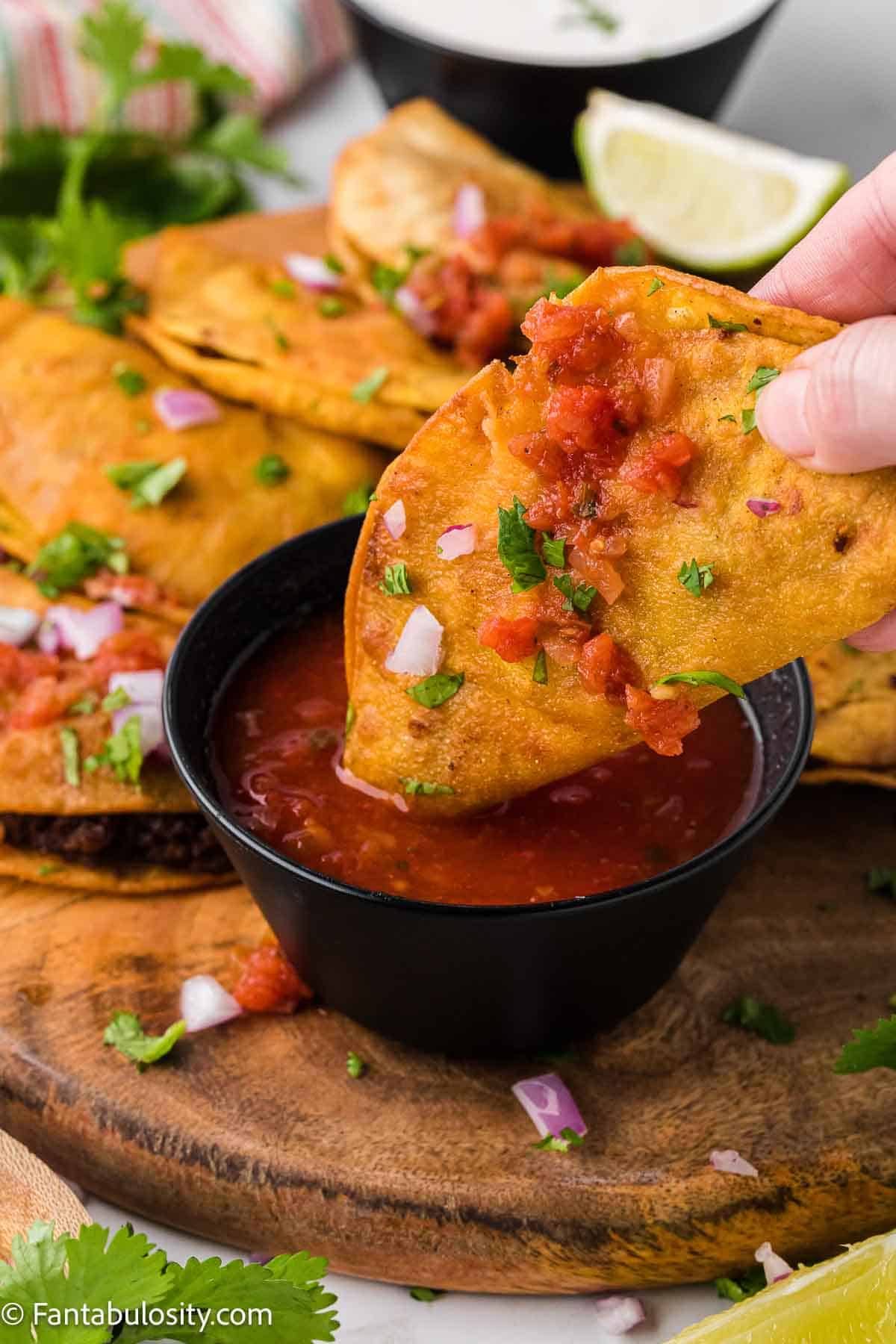 Fried taco being dipped in to salsa, on wooden cutting board.