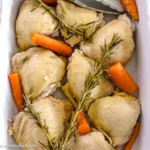 Boiled bone-in chicken thighs in white baking dish with rosemary and carrots.
