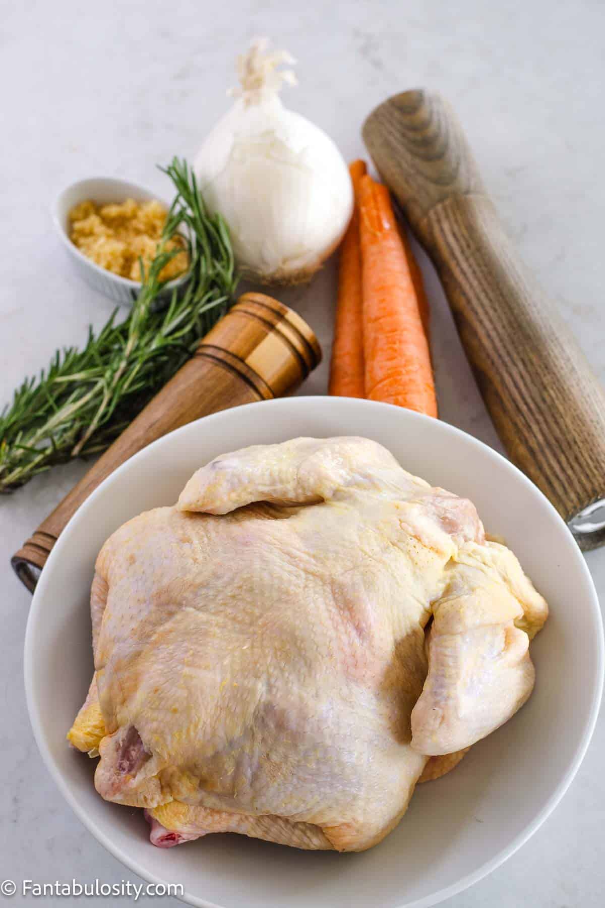 Raw, whole chicken, in a bowl, sitting next to salt and pepper, garlic, carrots, onion and fresh rosemary.