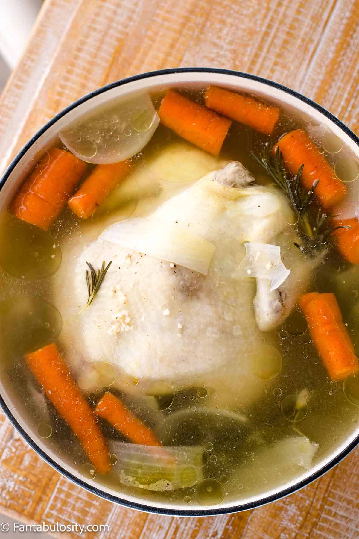 Cooked whole chicken in a dutch oven, surrounded by broth, and vegetables.