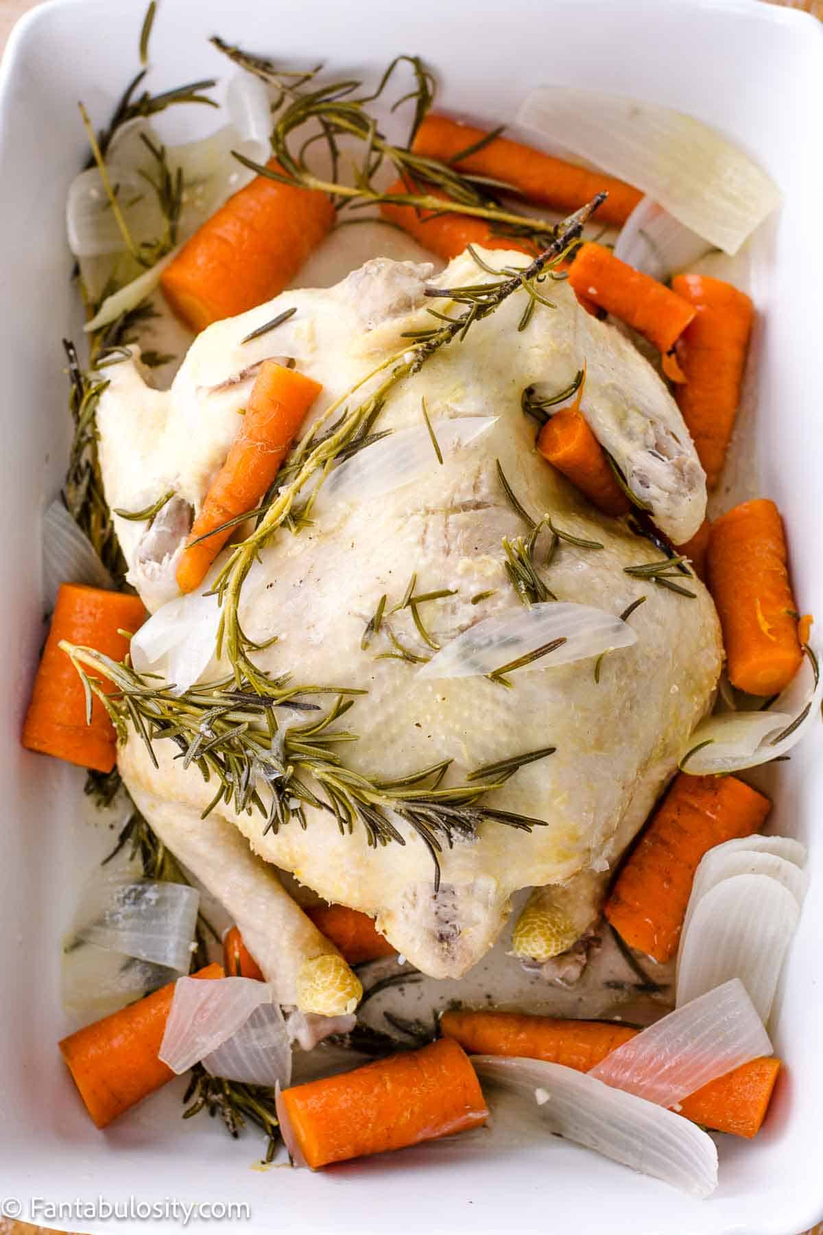 Cooked whole chicken in white dish, surrounded by vegetables and herbs.
