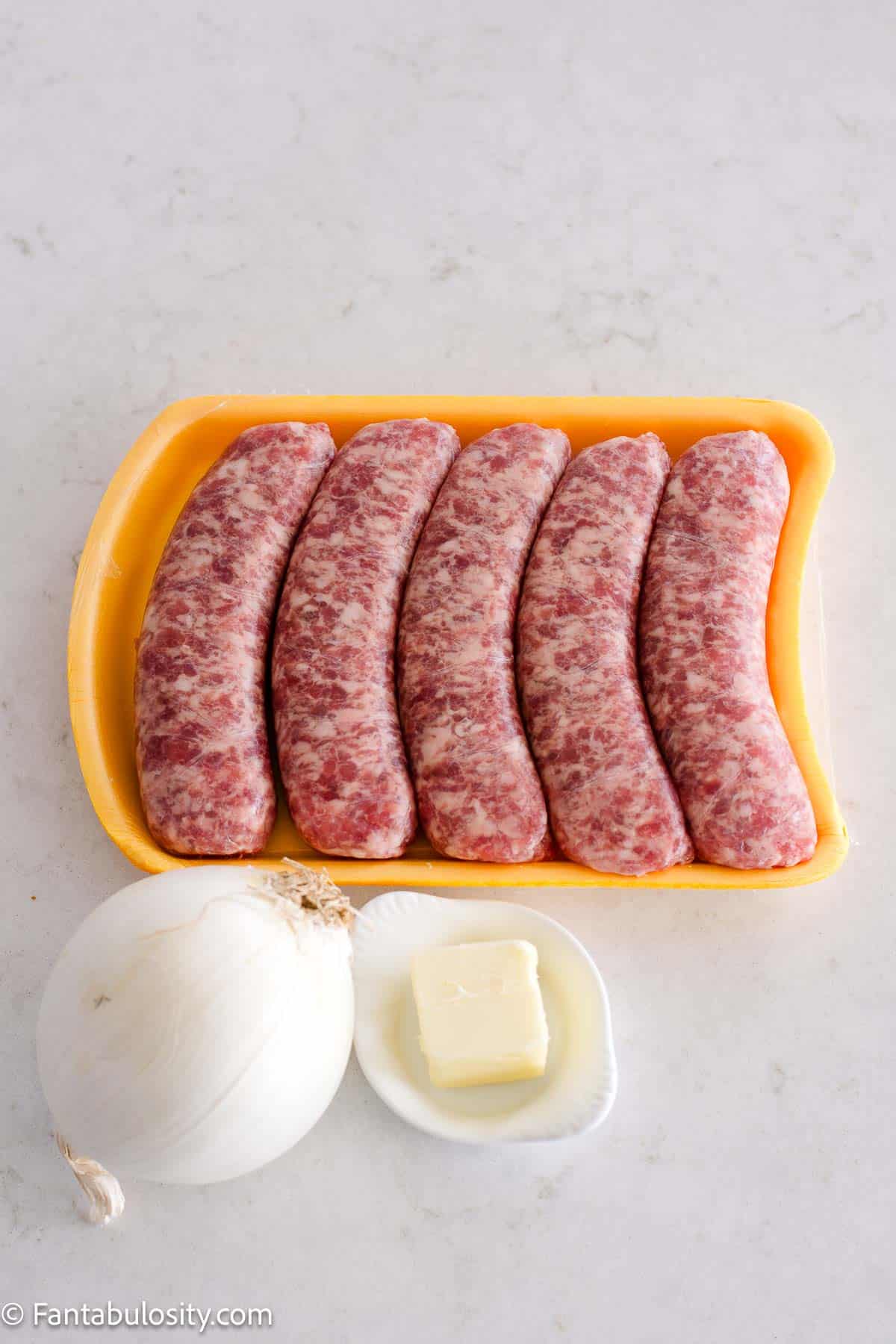 Raw, uncooked brats, sitting next to white onion and butter.
