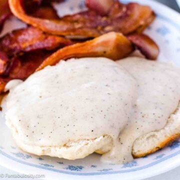 White gravy recipe on top of biscuits next to bacon on a vintage plate.