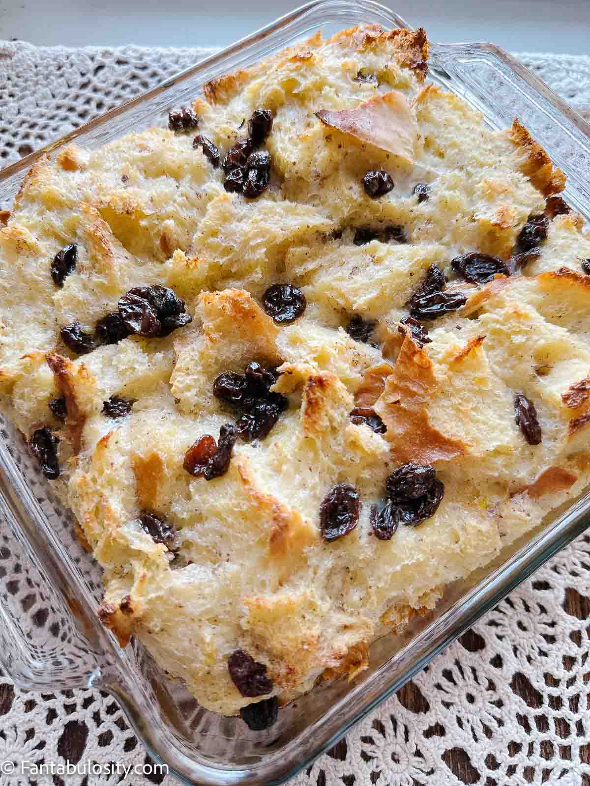 Bread pudding with raisins in baking dish, sitting on table, on top of lace cloth.
