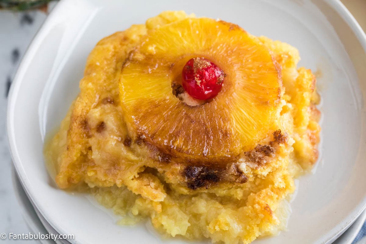 A serving of pineapple dump cake on a round plate, topped with pineapple ring and cherry