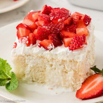 Slice of Strawberry Whipped Cream Cake on round plate, next to a sliced strawberry.