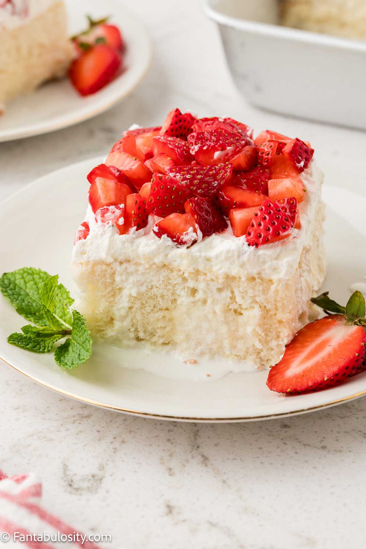 A slice of strawberry whipped cream cake on a round plate, with a piece of a mint leaf and sliced strawberry beside it.
