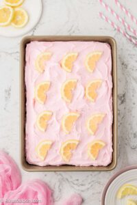 Pink lemonade cake in cake pan, with lemon slices on top of pink lemonade frosting.