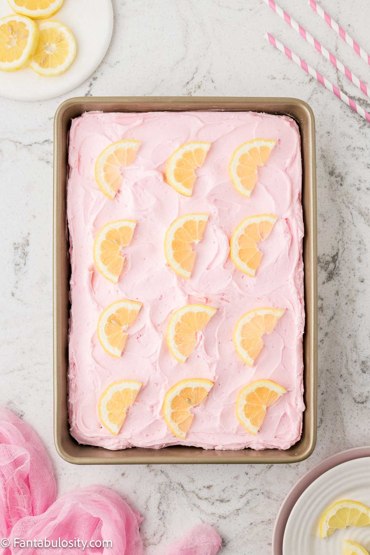 Pink lemonade cake in cake pan, with lemon slices on top of pink lemonade frosting.
