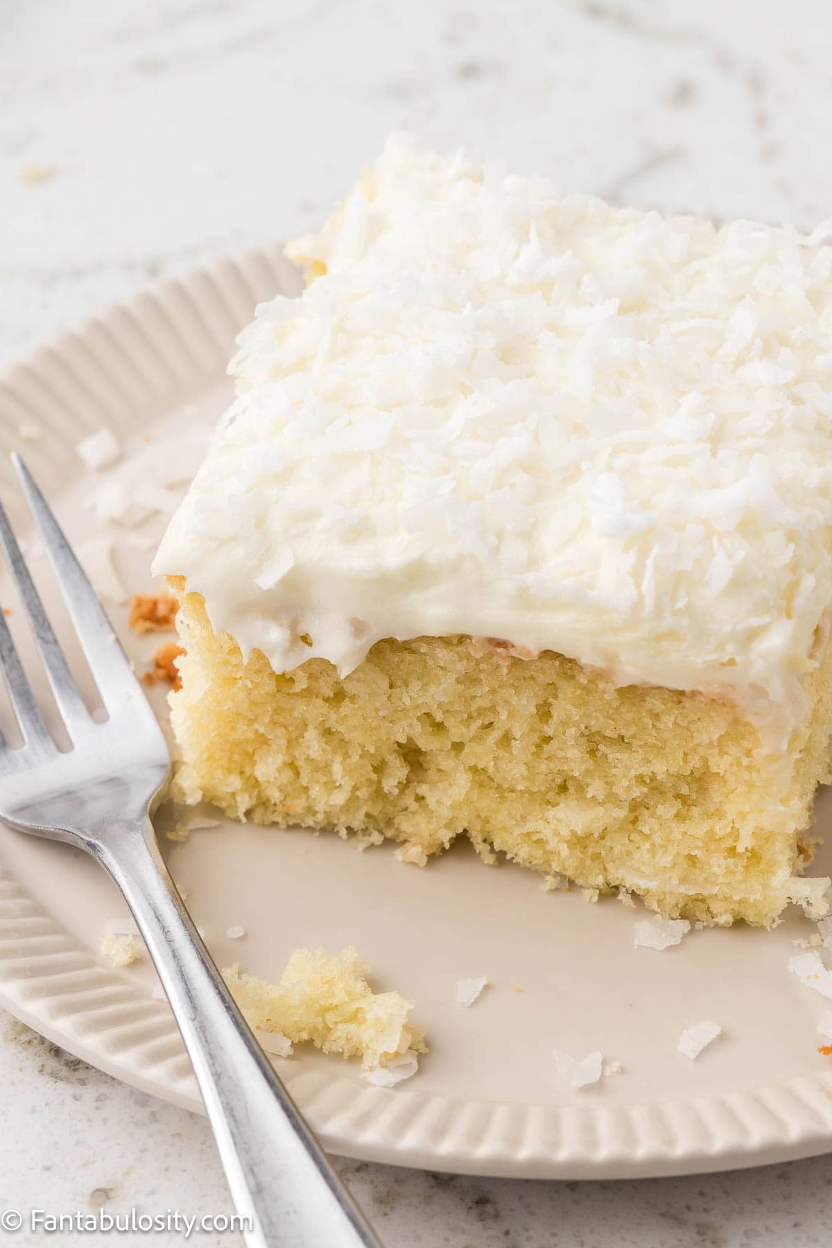 A piece of coconut sheet cake on a round plate, sitting next to a fork.
