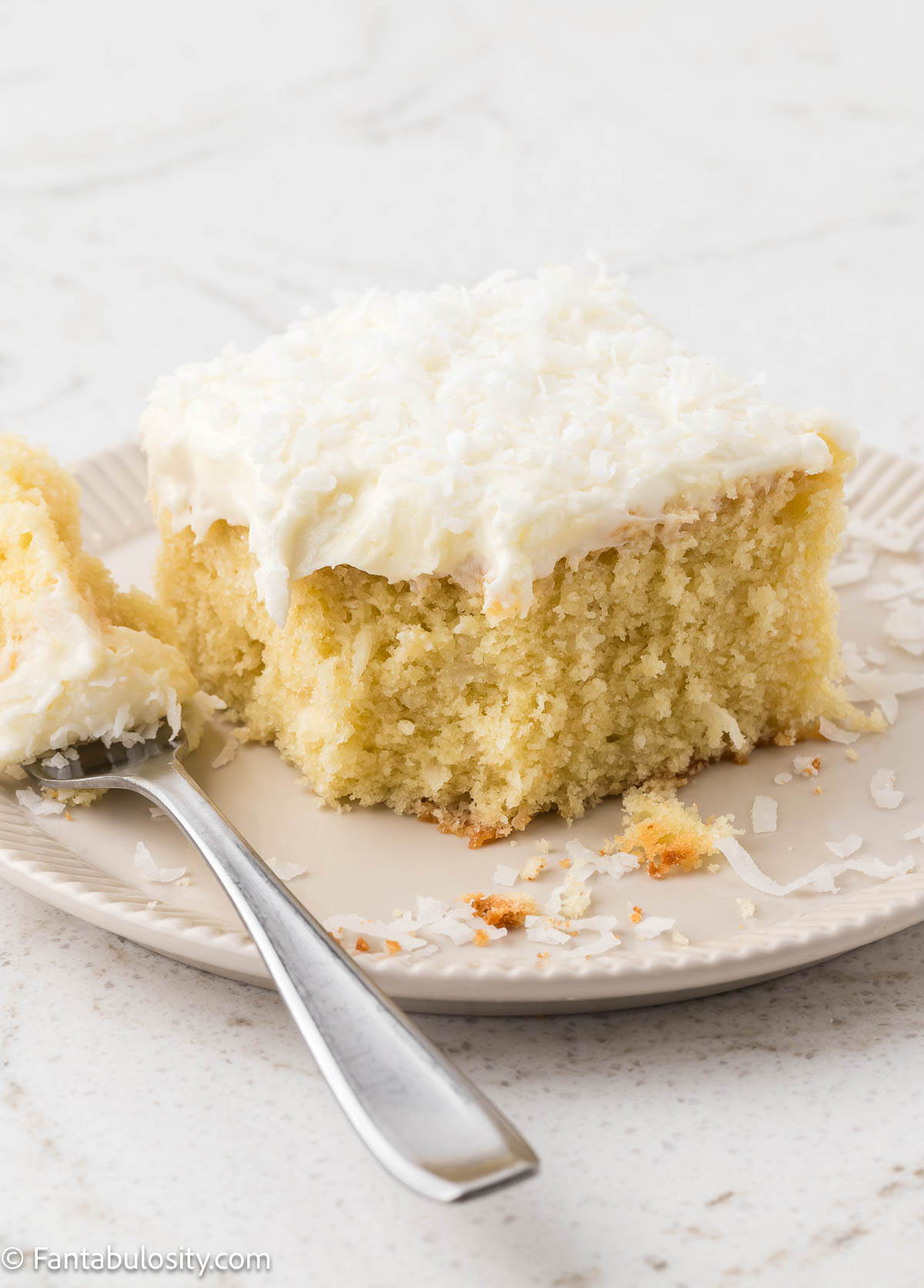 Slice of coconut cake on a round plate, with a fork sitting next to the cake with a bite on it.