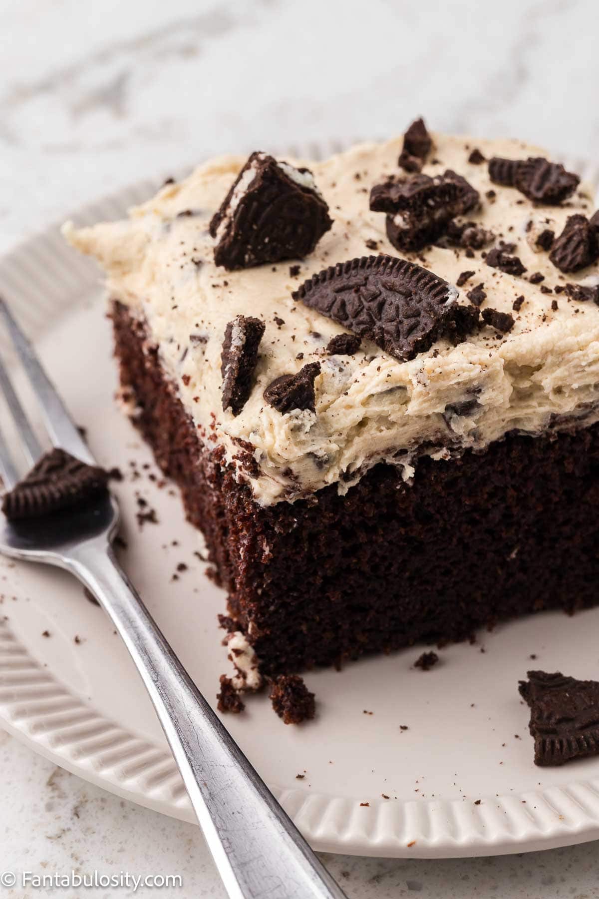 Slice of chocolate cake topped with Oreo cookie dough frosting, sitting on a round plate with a fork sitting next to the cake.
