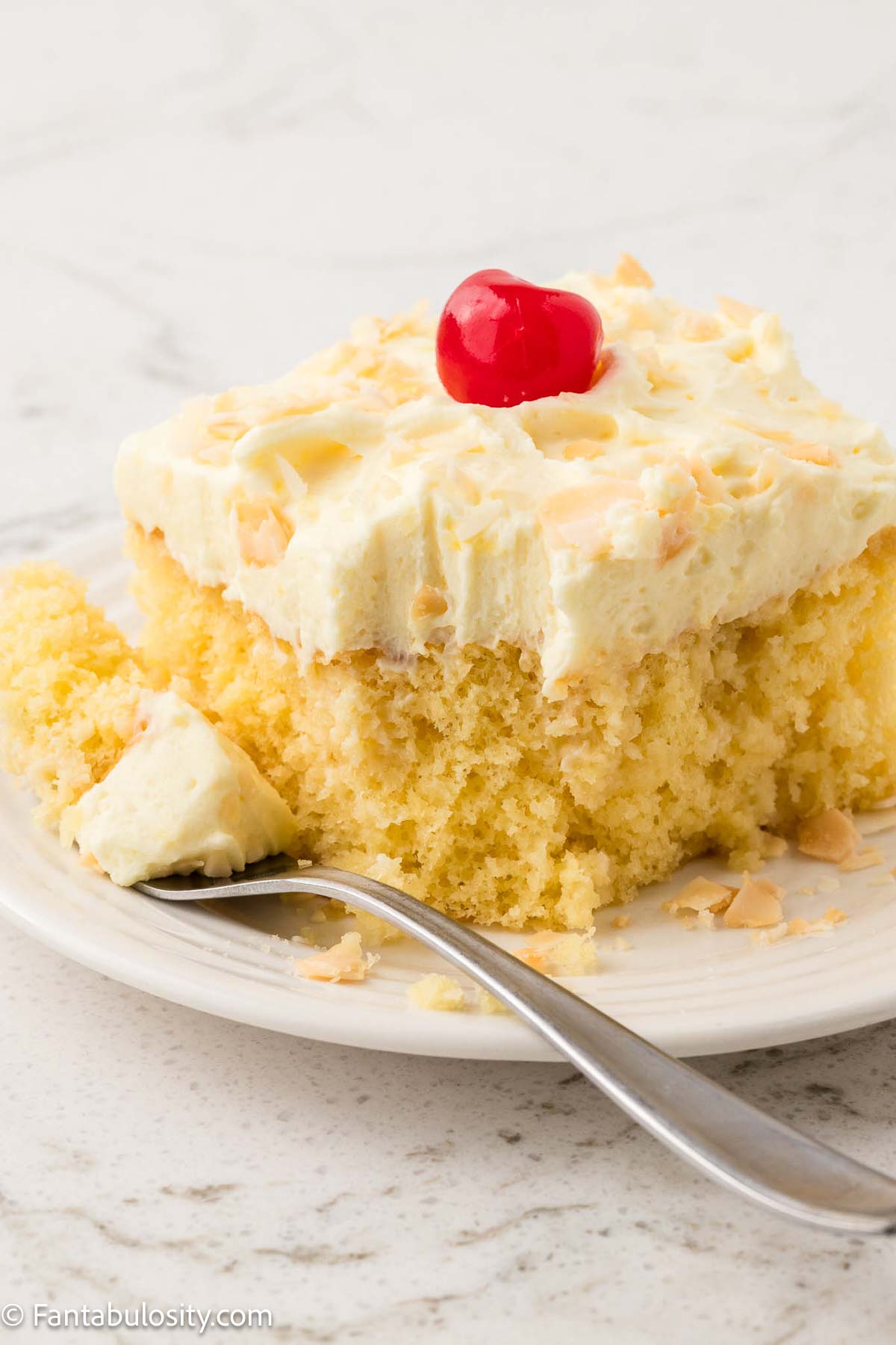 Slice of pina colada cake, topped with frosting, and a cherry, sitting on a round plate, with a fork lying next to it.