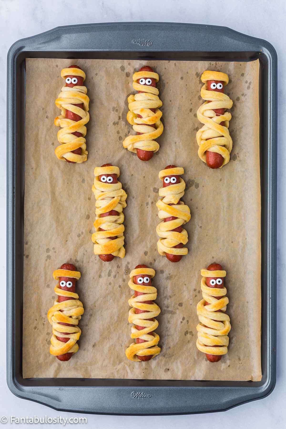 Mummy hot dogs with candy eyes, sitting on baking pan.