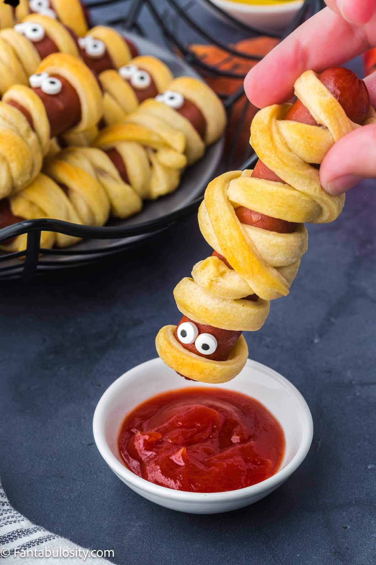 Mummy hot dog being dipped in to small bowl of ketchup.