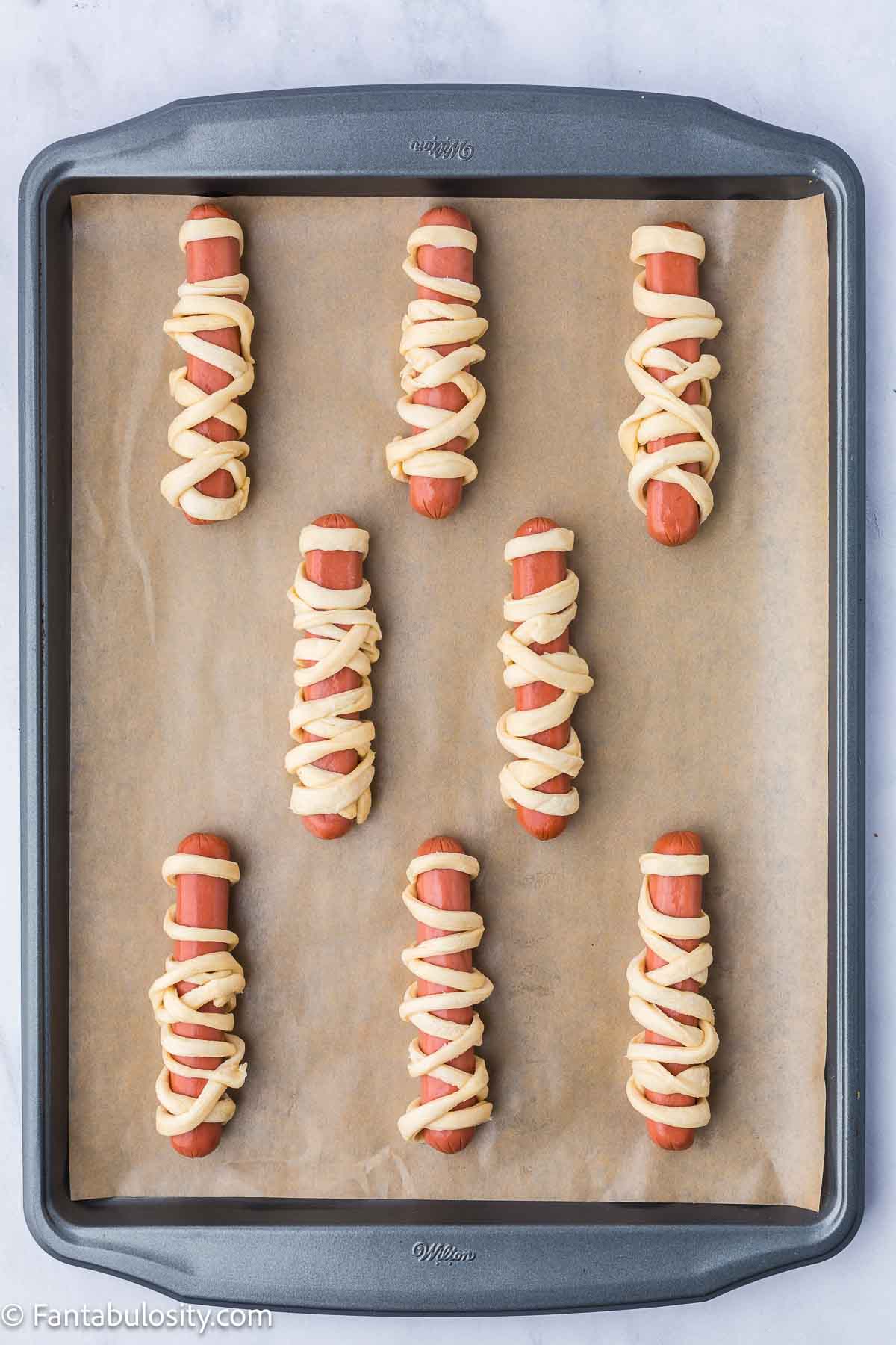 Hot dogs wrapped in crescent roll strips, made to look like mummies, resting on a baking pan.
