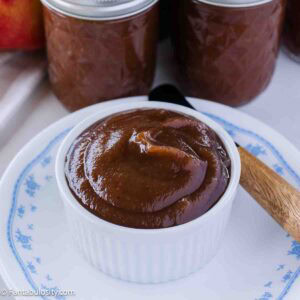 Smooth slow cooker apple butter in small bowl, sitting next to canned jars of apple butter.