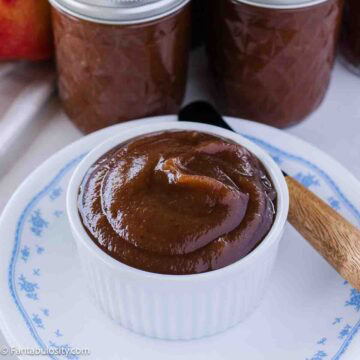Smooth slow cooker apple butter in small bowl, sitting next to canned jars of apple butter.