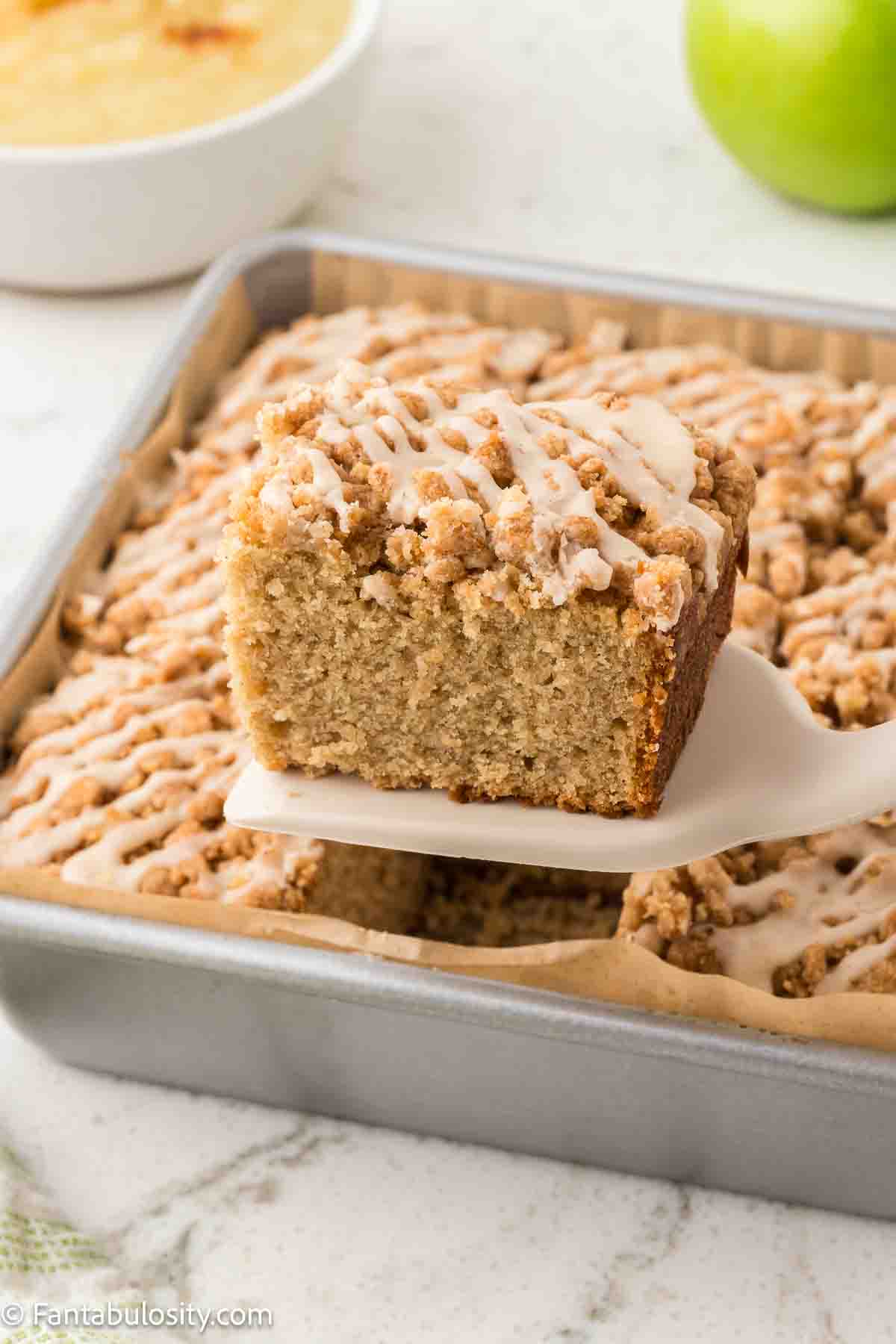 Slice of applesauce crumb cake on spatula, resting above rest of cake.