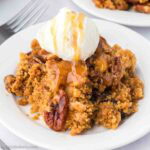 One serving of pecan pie dump cake on a plate topped with vanilla ice cream and caramel drizzle.
