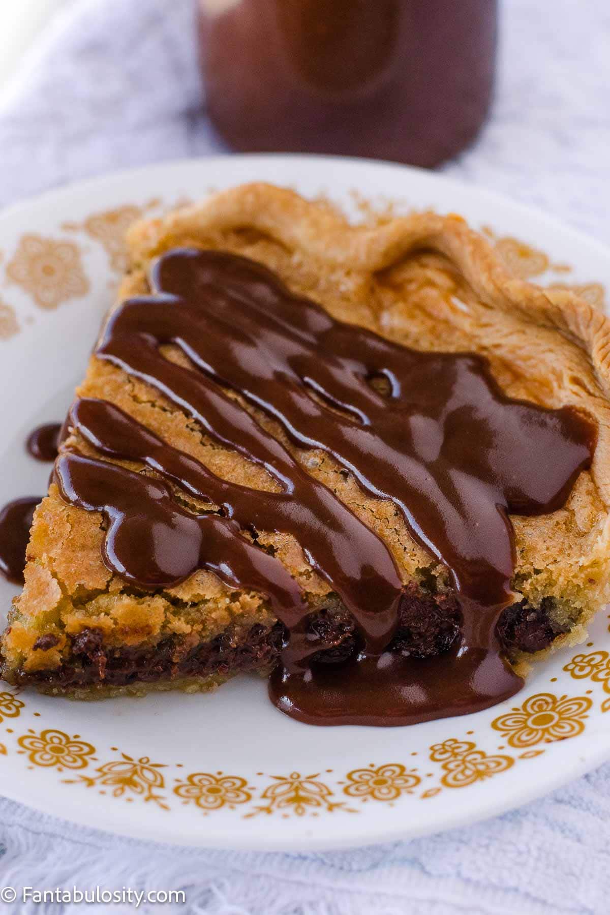A slice of chocolate chip cookie pie on plate that's topped with hot fudge sauce.