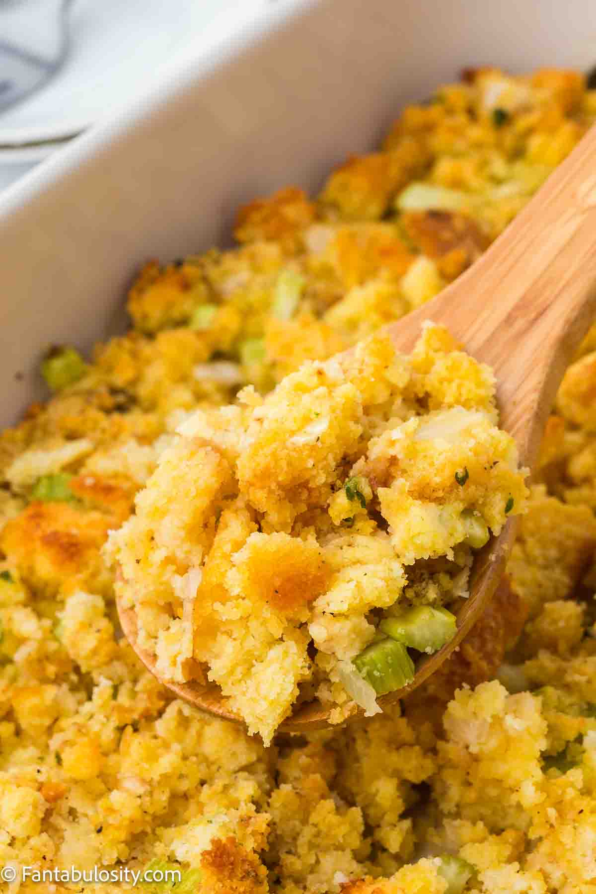 Cornbread dressing being scooped up out of baking dish with wooden spoon.