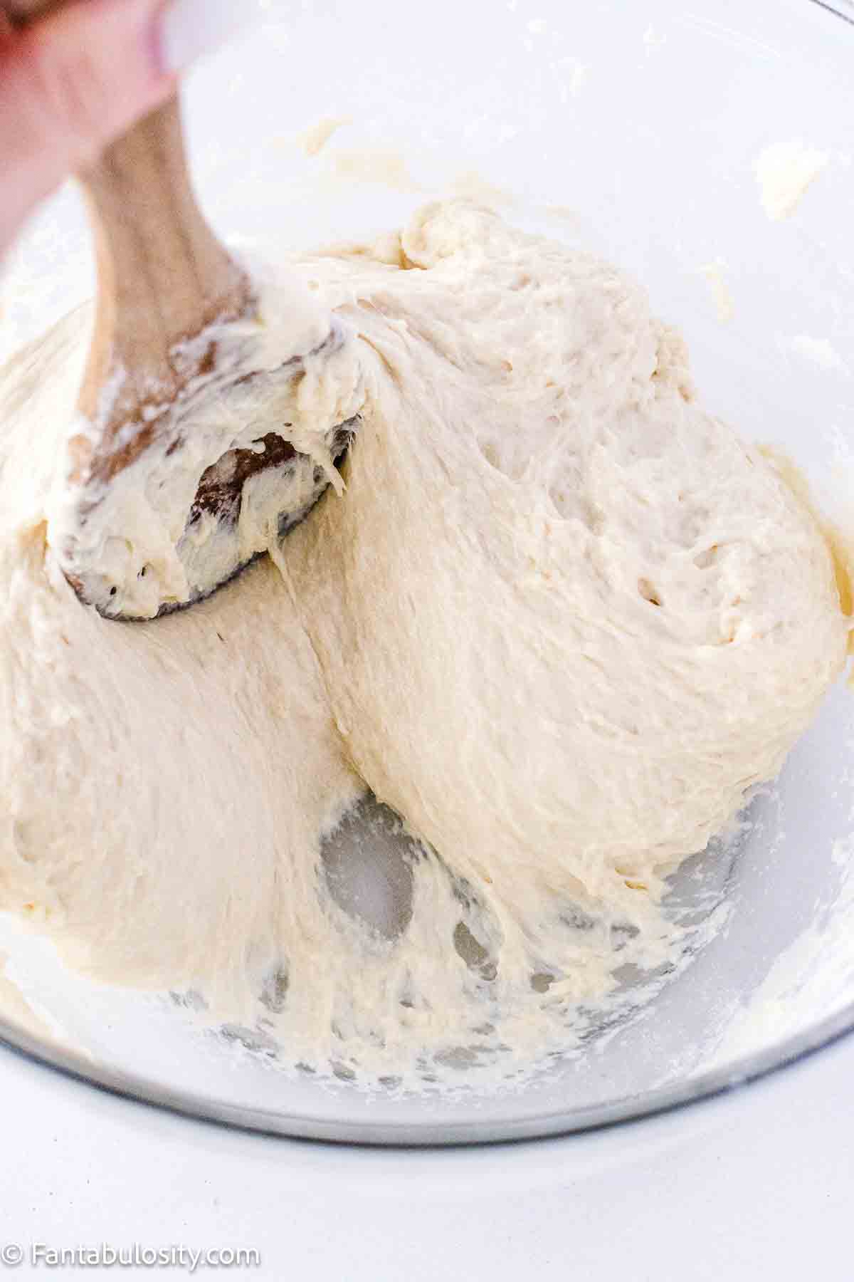 Wooden spoon pulling roll dough away from the sides of the mixing bowl, showing that it's ready.