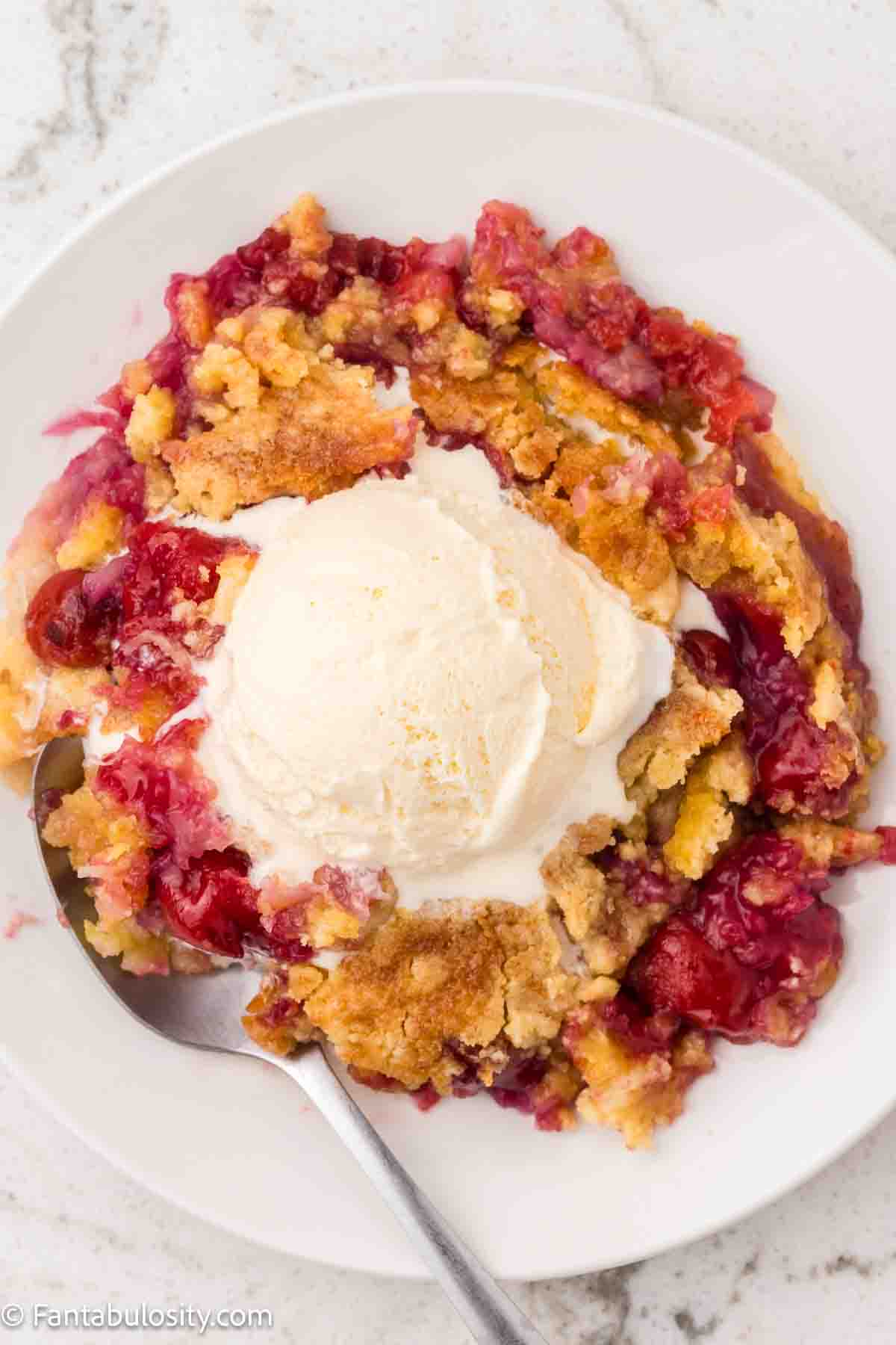 A serving of pineapple cherry dump cake on round plate, topped with vanilla ice cream scoop.