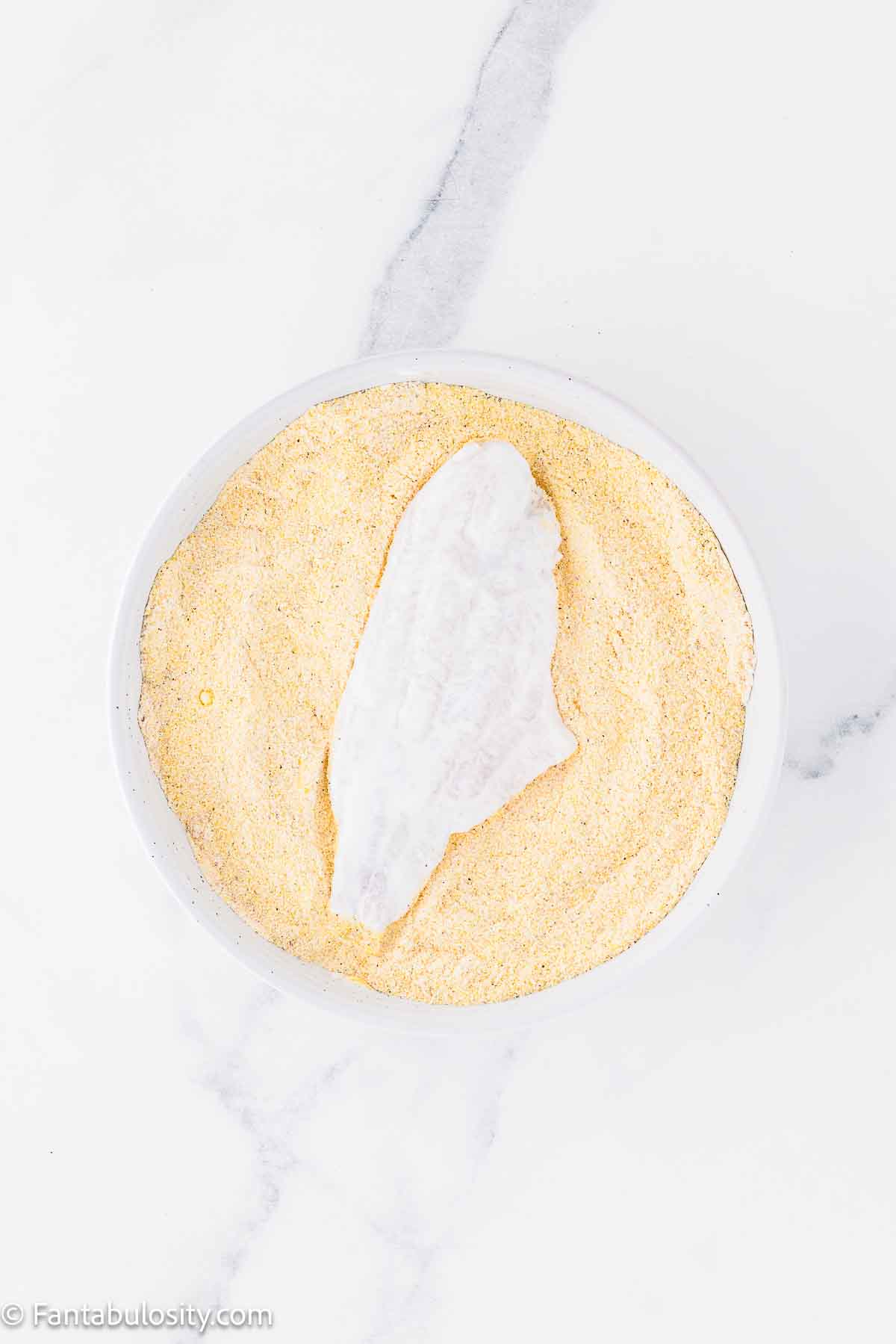 Buttermilk soaked catfish dipped in flour and seasoning mixture in shallow dish.