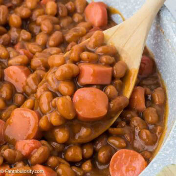 A pan full of beanie weenies with a wooden spoon dishing out a serving.